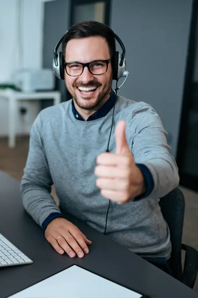Smiling Caucasian man with glasses and mustache giving thumbs up while wearing headphones with microphone in front of computer, demonstrating successful small business review management.