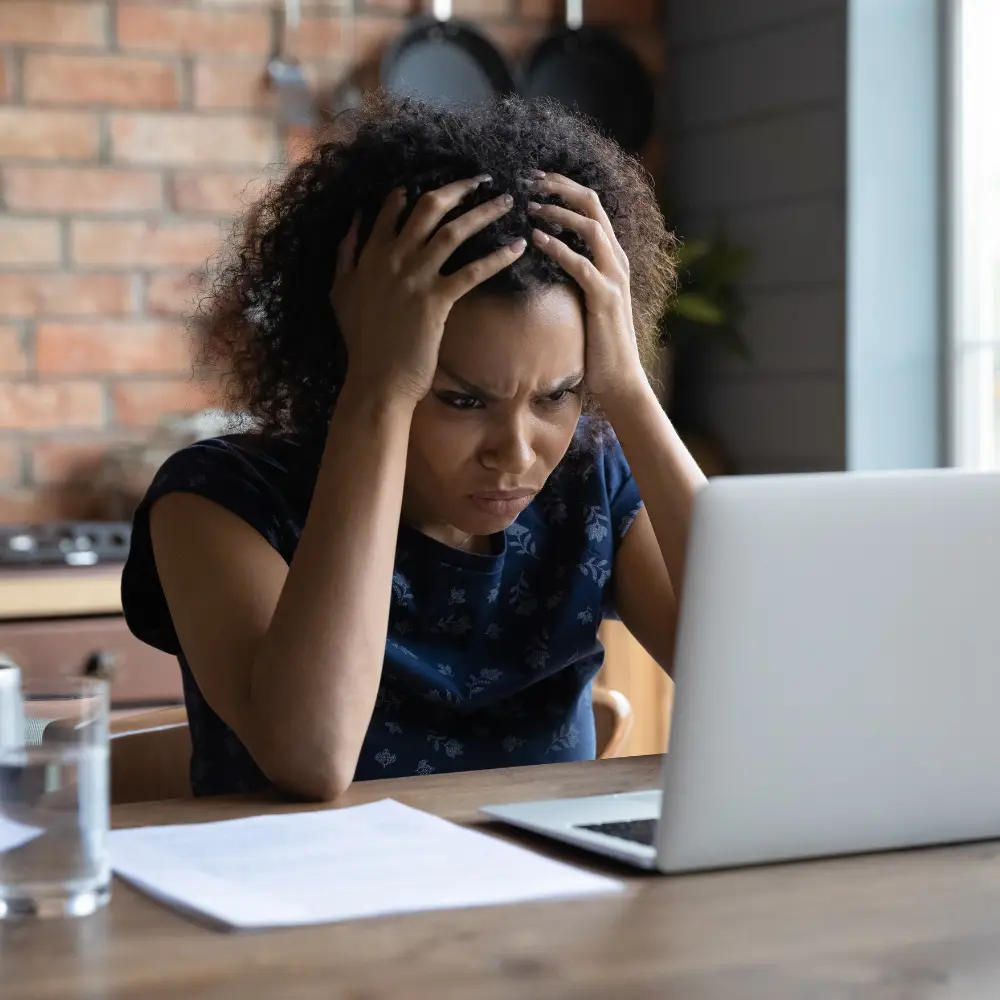 A frustrated woman sitting at a table with both hands on her head, elbows resting on the table, overwhelmed by small business reputation management tasks. An open laptop is in front of her with a white sheet of paper to the right. The background features a brick wall with hanging pans and a small kitchenette, conveying a cozy yet stressed atmosphere. The image highlights the challenges faced in maintaining a positive small business reputation management strategy